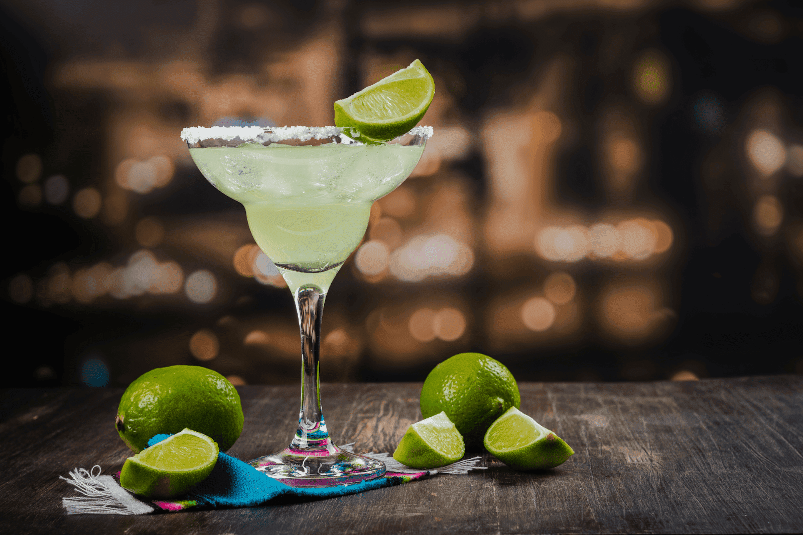 Classic margarita in a stemmed glass with a salted rim and lime wedge, surrounded by lime slices on a rustic wooden table
