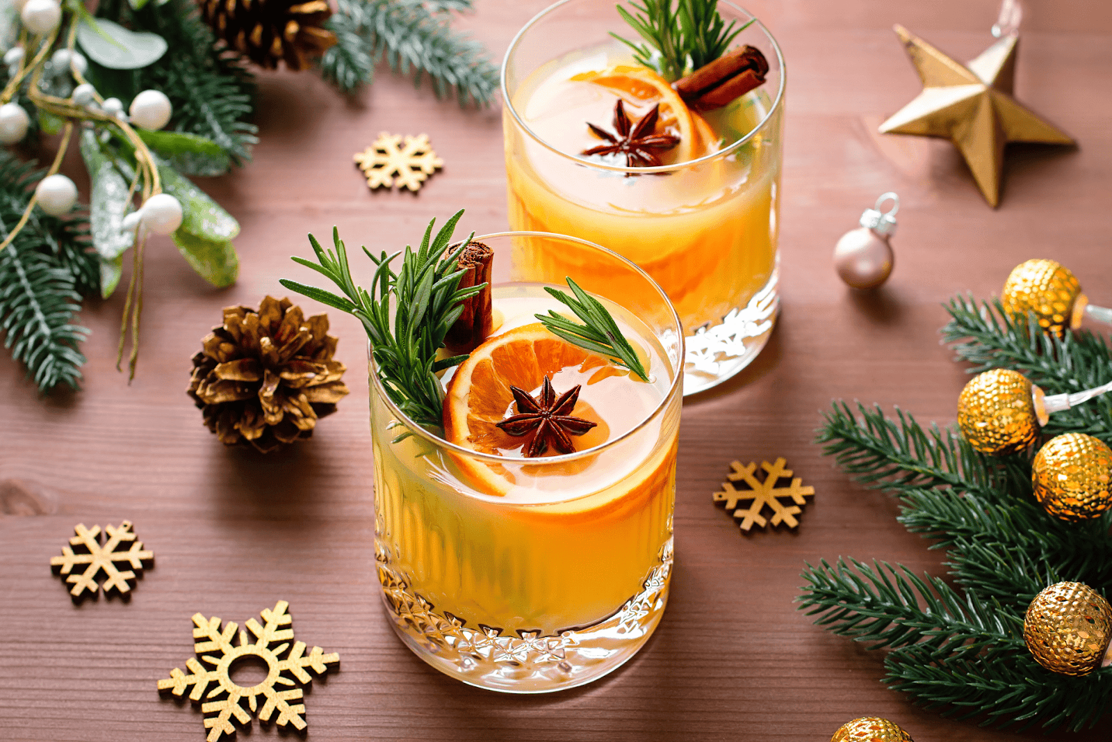 Holiday cocktails with orange slices, rosemary, and cinnamon sticks surrounded by festive decorations.