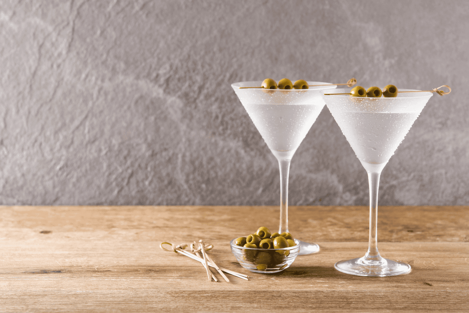 Two martini glasses with olives on a wooden table against a gray background