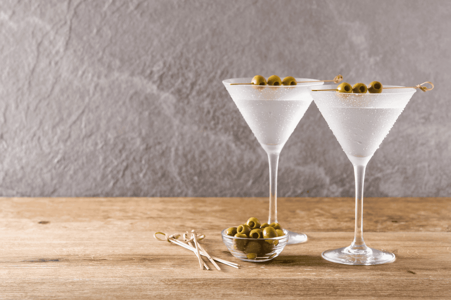 Two martini glasses with olives on a wooden table against a gray background