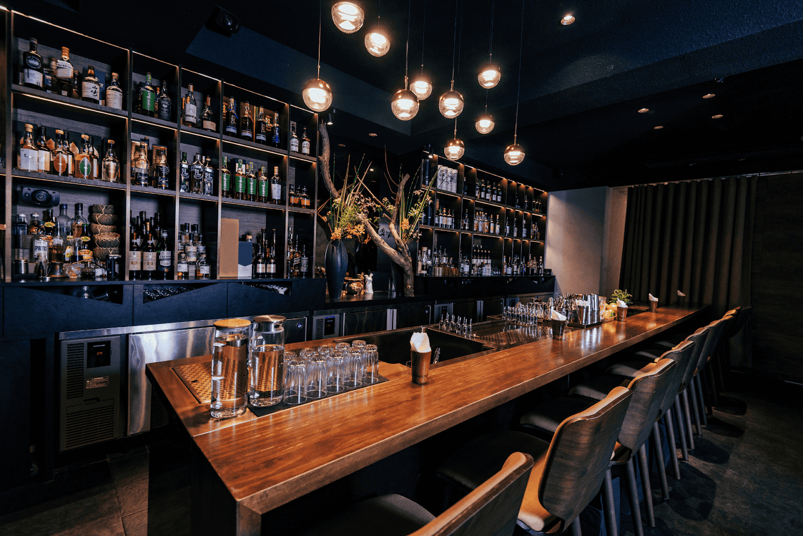 Modern bar interior with wooden counter, shelves of liquor bottles, and warm lighting.