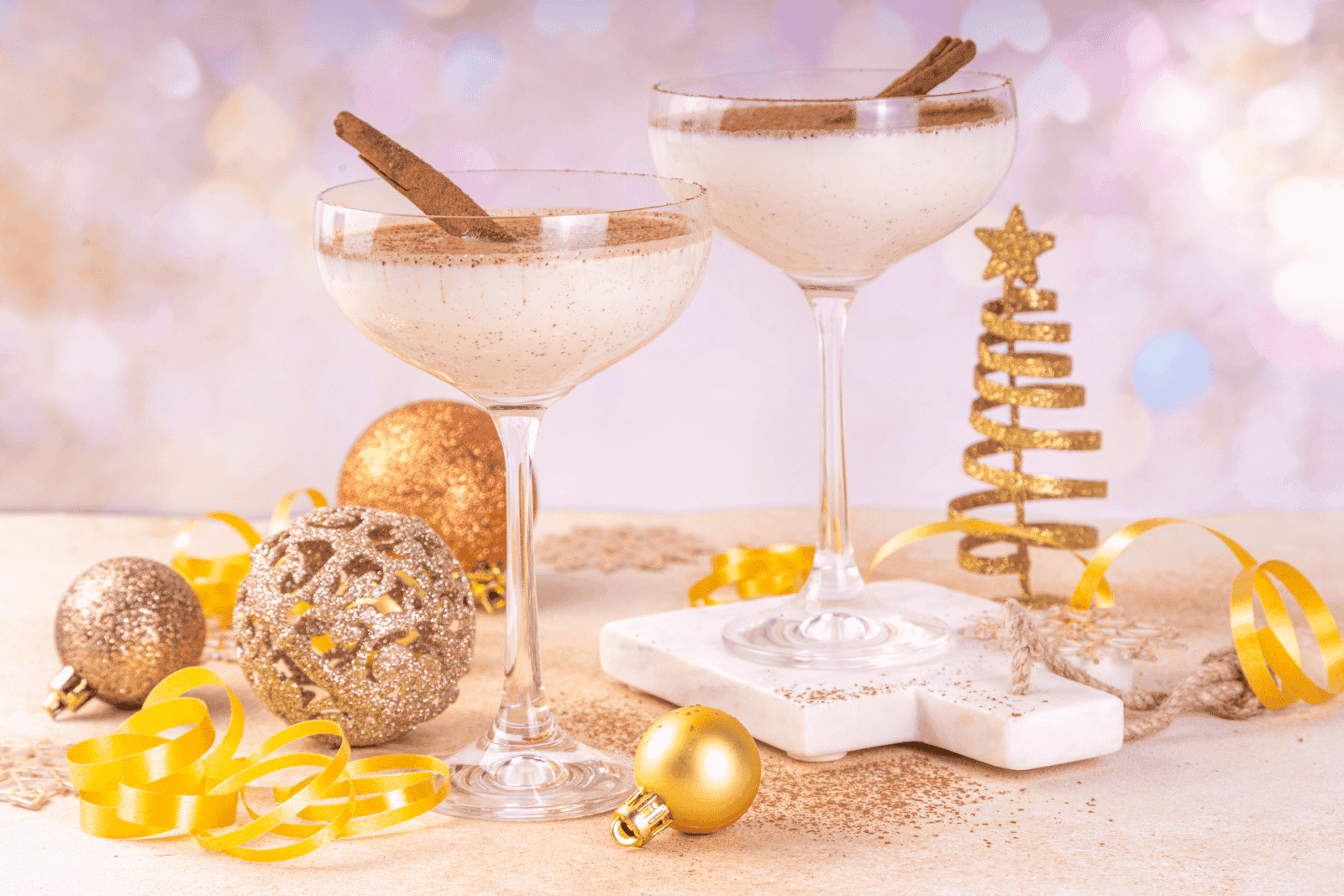 Two creamy Eggnog Martinis in coupe glasses with cinnamon sticks, surrounded by festive holiday decorations