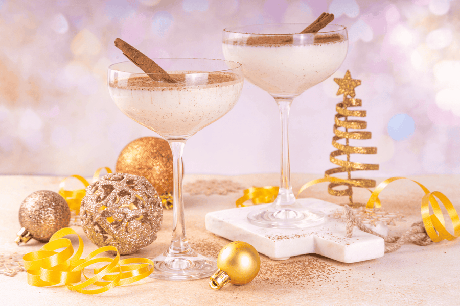 Two creamy Eggnog Martinis in coupe glasses with cinnamon sticks, surrounded by festive holiday decorations