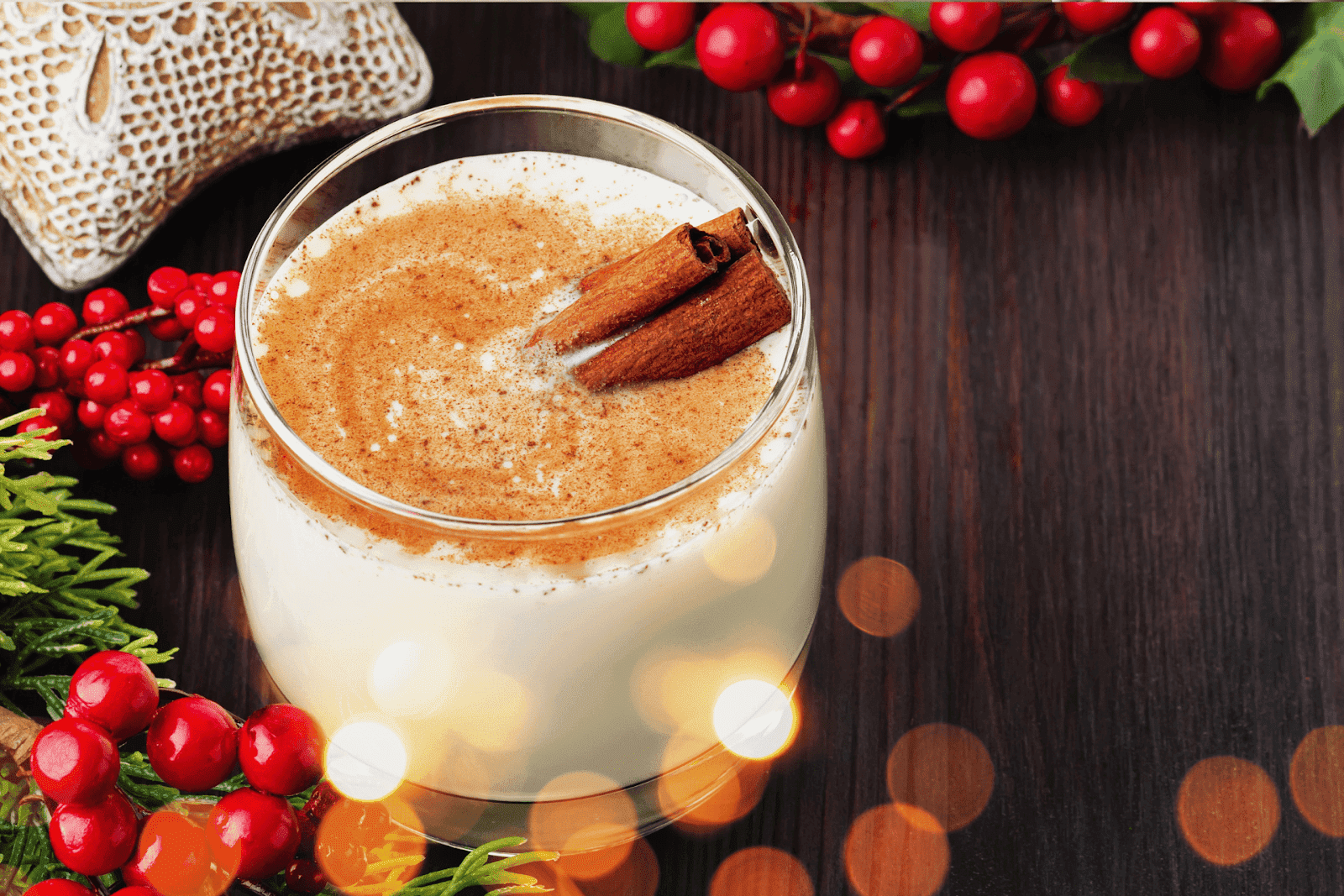 Glass of creamy eggnog topped with ground cinnamon and cinnamon sticks, surrounded by holiday greenery and red berries