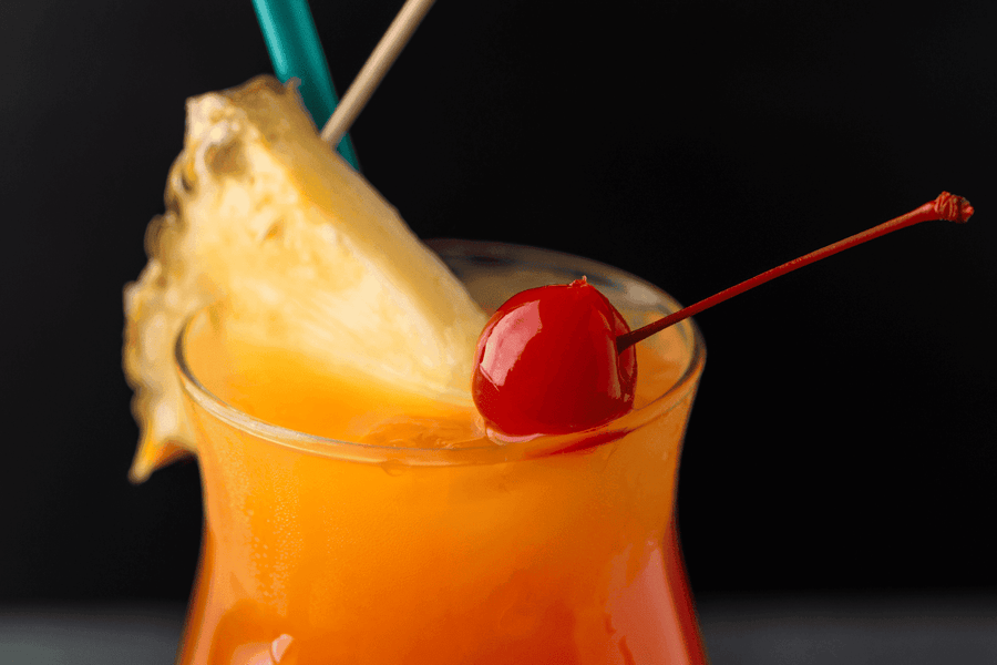 Tropical cocktail with a pineapple wedge and maraschino cherry garnish, showing vibrant orange and yellow layers in a hurricane glass