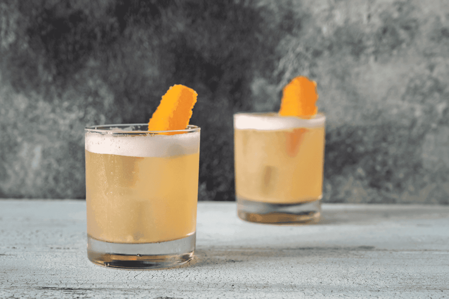Two whiskey sour cocktails with frothy tops and orange peel garnish