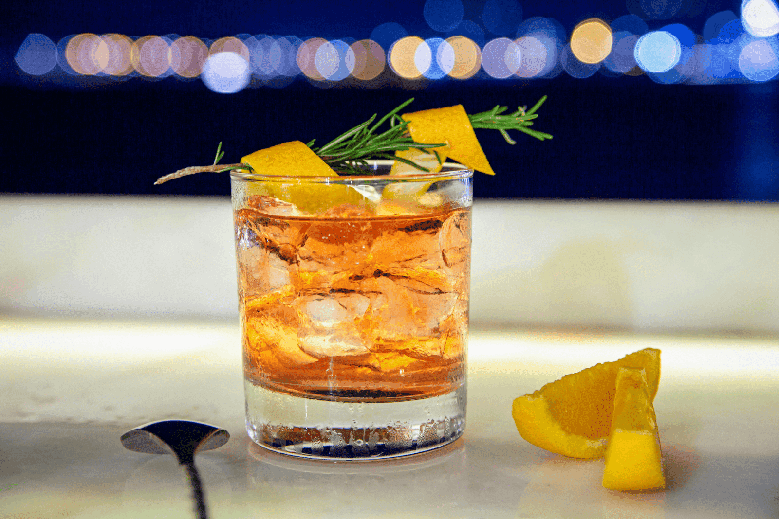 Glass of Old Fashioned cocktail with ice, garnished with an orange peel twist and rosemary, set against blurred night city lights