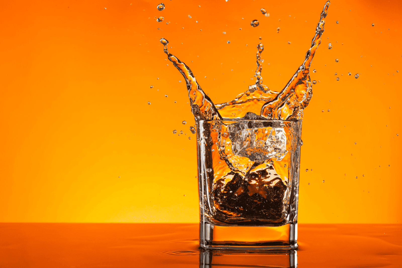 Ice cubes splashing into a glass of whiskey with an orange background.
