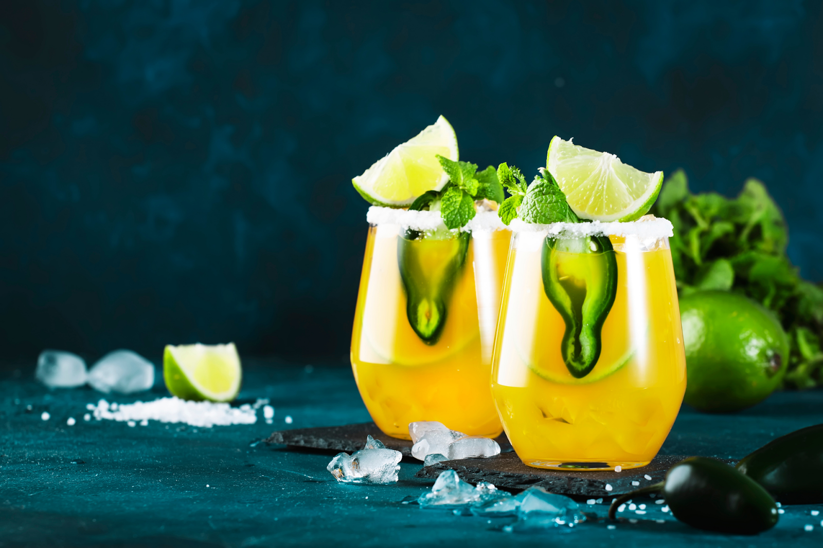 Bright spicy margaritas with lime wedges, jalapeño slices, and mint sprigs in glasses with salted rims on a dark background