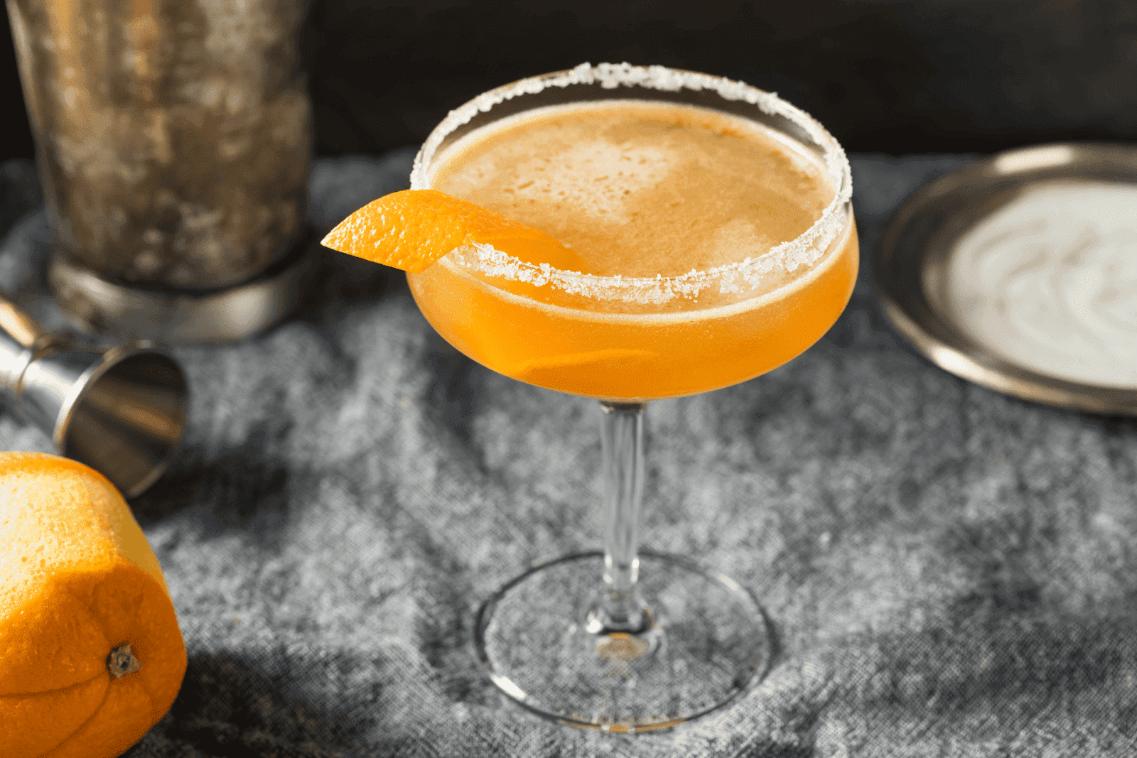 A Sidecar cocktail in a coupe glass with a sugared rim and orange garnish, set on a textured surface