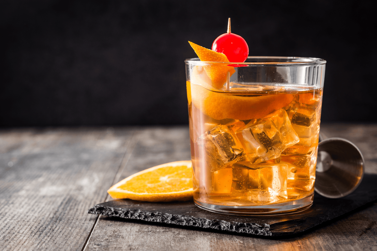 Old Fashioned cocktail with orange peel, cherry, and ice on a rustic wooden table.