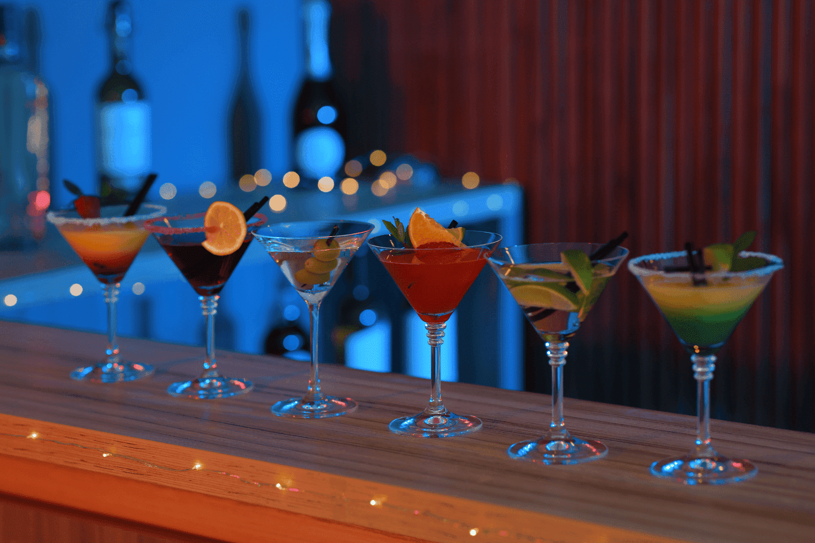 Variety of martini cocktails lined up on a bar with colorful garnishes and lights in the background