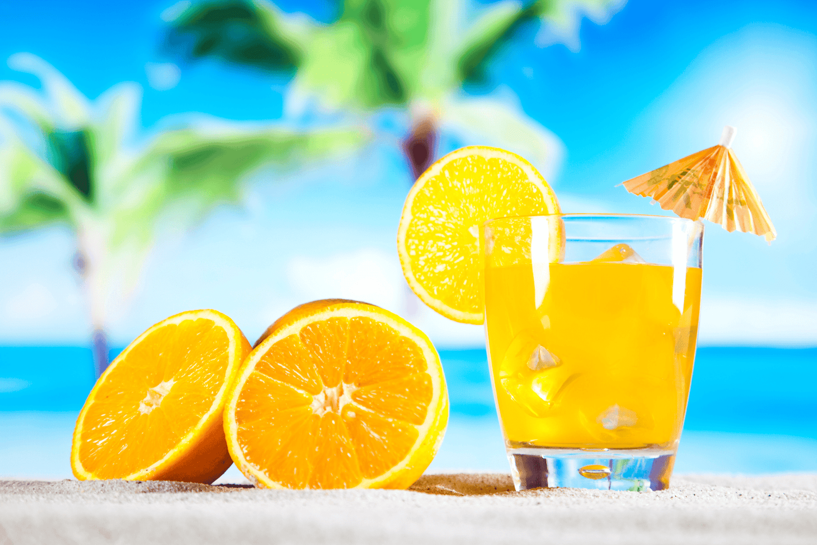 Glass of orange cocktail with ice and sliced oranges on a beach background.
