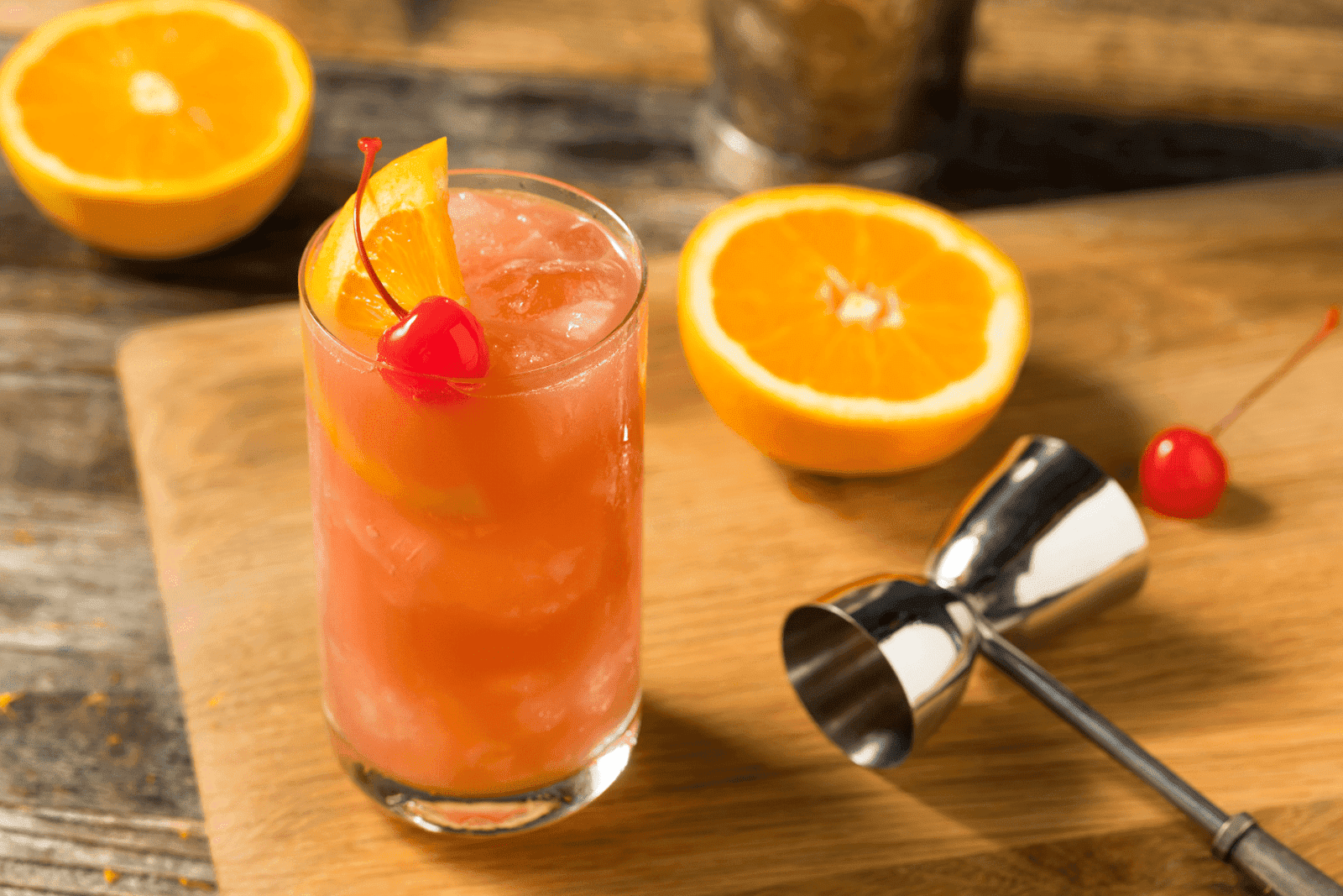 A Sex on the Beach cocktail on a wooden surface, garnished with an orange slice and cherry, surrounded by halved oranges and a metal jigger.