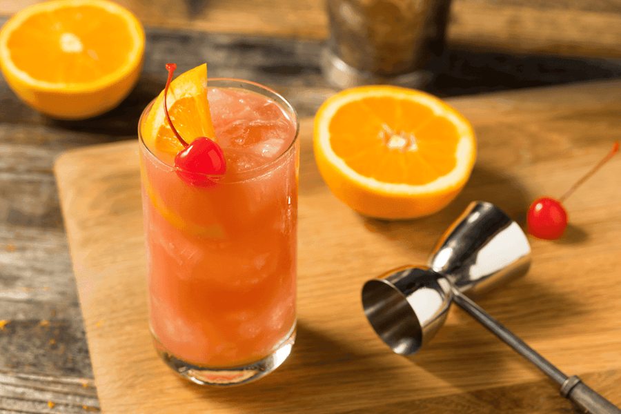 A Sex on the Beach cocktail on a wooden surface, garnished with an orange slice and cherry, surrounded by halved oranges and a metal jigger.