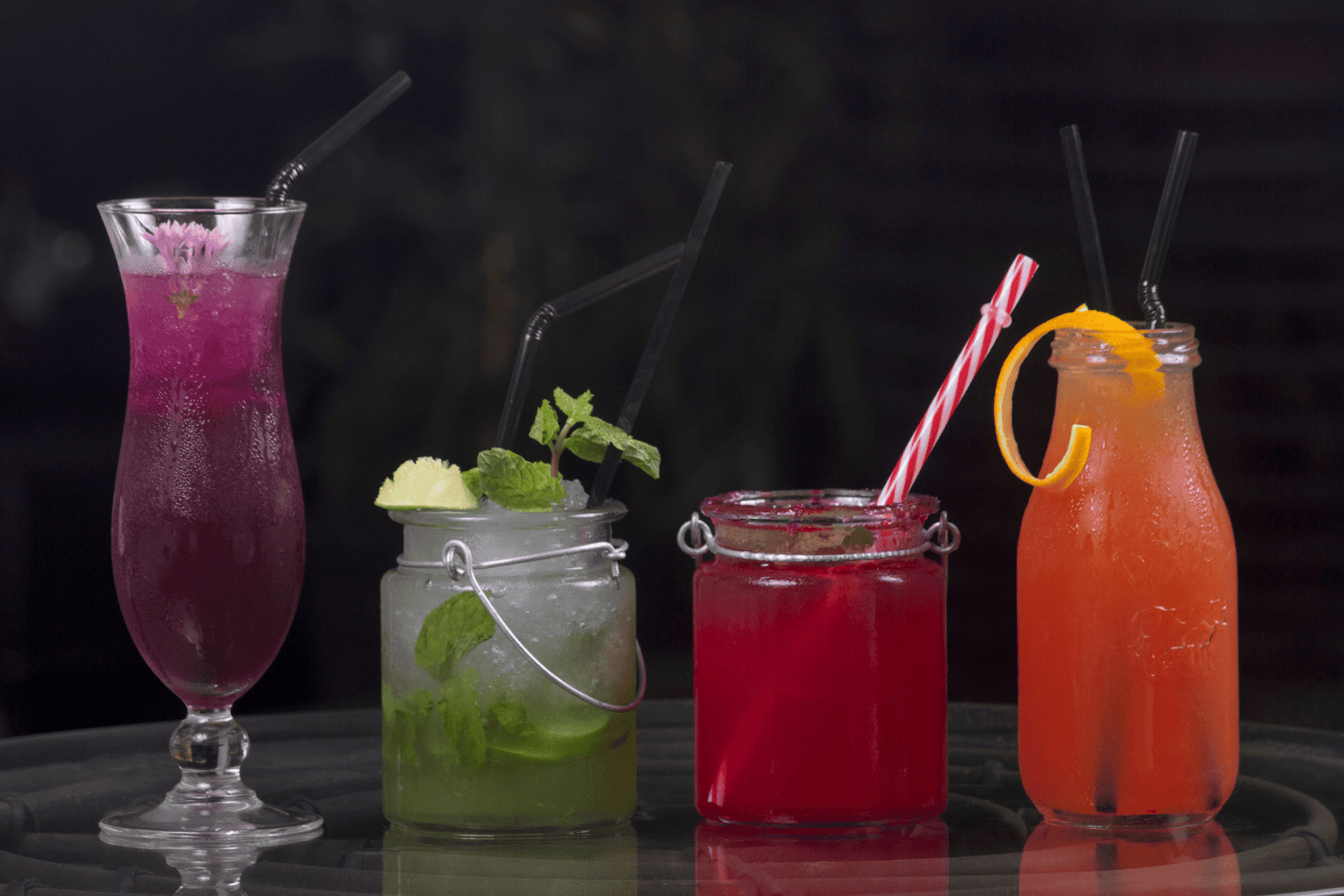 Four colorful mocktails in different glasses with straws, garnished with fruit and mint.