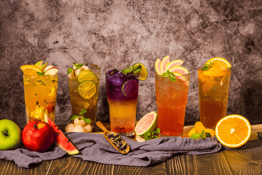 Five tall glasses of colorful fruit mocktails with citrus and mint garnishes on a rustic table.