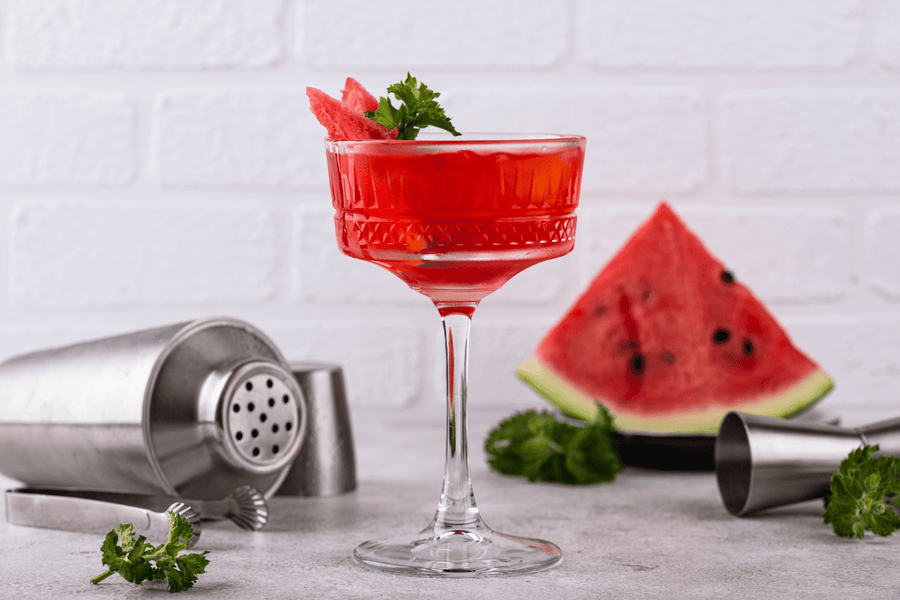 Red watermelon margarita mocktail in a glass garnished with mint and watermelon slice, with cocktail tools and fruit in the background