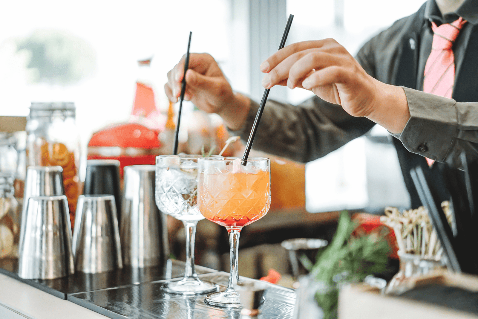 Bartender stirring two colorful cocktails with straws at a bar counter.