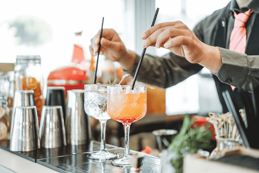 Bartender stirring two colorful cocktails with straws at a bar counter.