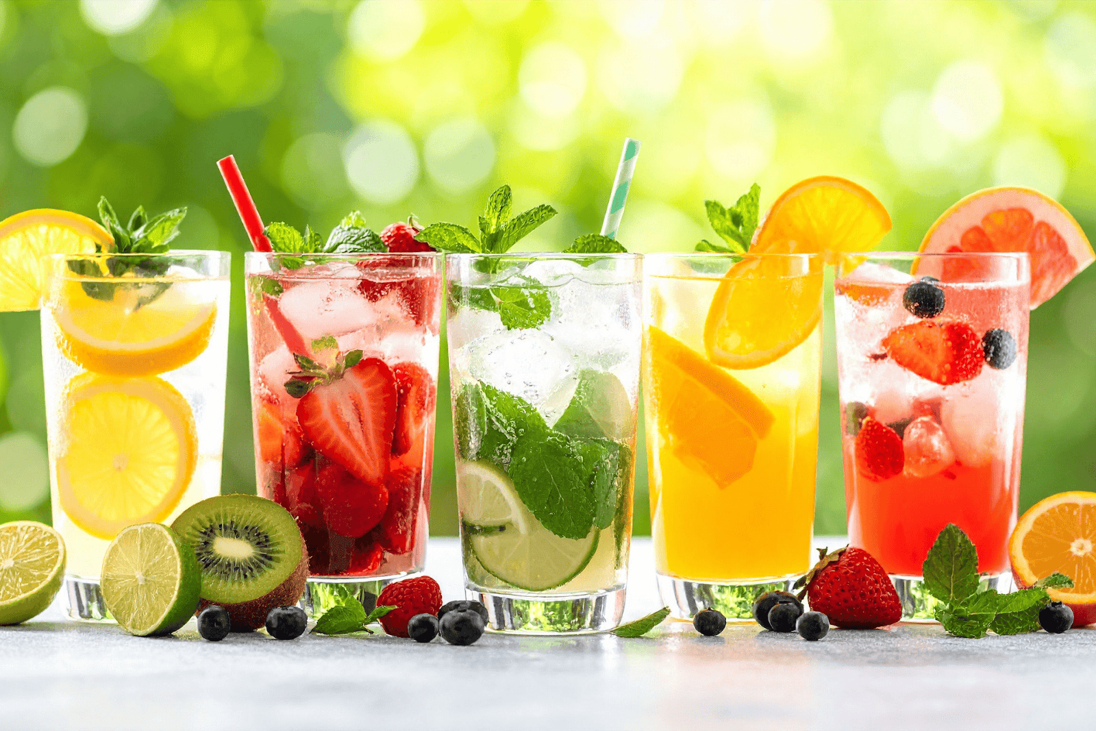 Fresh fruit drinks with ice, citrus slices, and berries in glasses on a bright outdoor background.