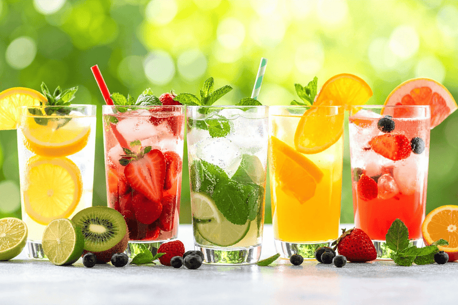 Fresh fruit drinks with ice, citrus slices, and berries in glasses on a bright outdoor background.