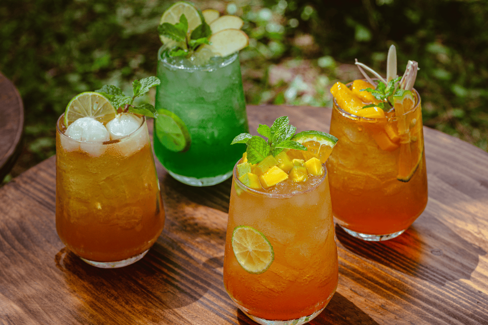 Four tropical fruit mocktails with mint and citrus garnishes on a wooden table outdoors.