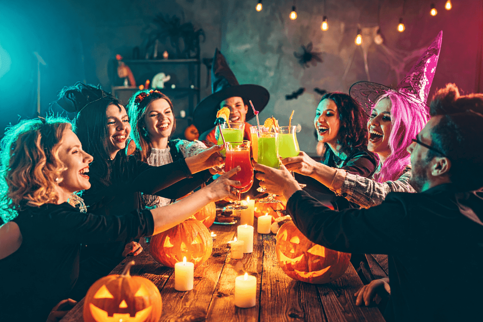 Group of friends in Halloween costumes raising colorful drinks surrounded by pumpkins and candles.
