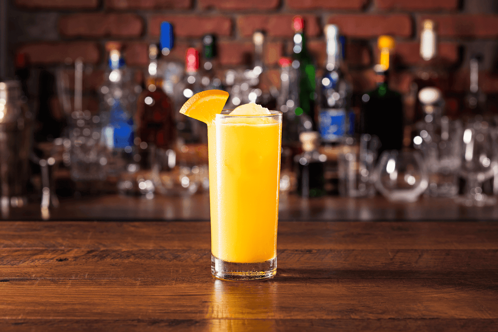 A tall glass of Screwdriver cocktail with ice and an orange wedge garnish, placed on a bar counter with bottles blurred in the background.