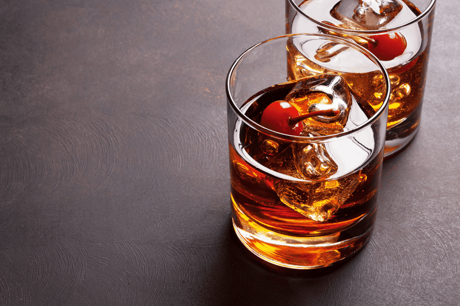 Two Manhattan cocktails on the rocks, garnished with maraschino cherries and ice cubes.